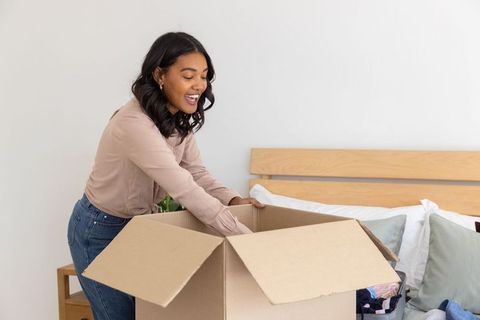 African american woman unpacking moving box in minimalist bedroom smiling