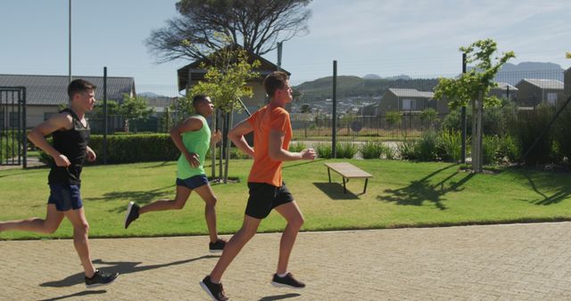 Three men are running in the open, wearing sports clothes, on a bright sunny day. Houses and greenery are in the background, suggesting a residential area or park. This image might be suitable for uses related to fitness, exercising, or lifestyle blogs. It can also be used in advertisements for sportswear, fitness programs, or community health initiatives.