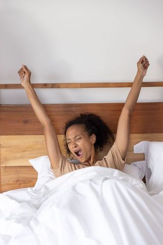 African american woman stretching and yawning in cozy bed with wooden headboard and white duvet