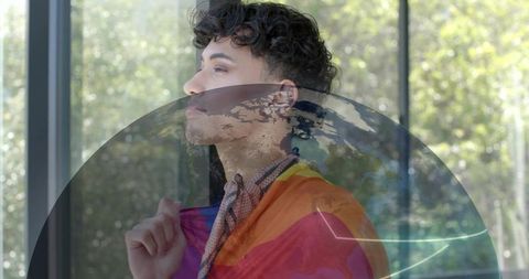 Contemplative man standing with rainbow flag and globe reflection in sunlit studio window