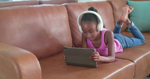 Teen girl lying on leather sofa using tablet and headphones for online learning at home