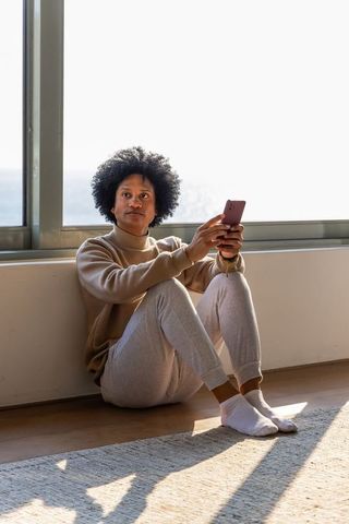 Sunlit african american man sitting by window using smartphone in minimal home interior
