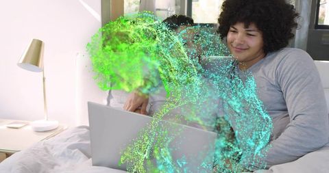 Two men watching laptop on bed in sunlit bedroom with teal-green digital particle overlay