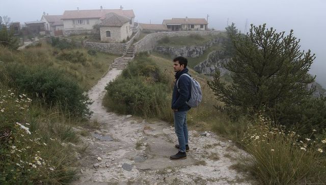 Solo hiker standing on foggy cliff trail overlooking stone cottages on rural mountain path