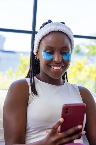 African american woman wearing blue hydrogel under-eye patches smiling while using phone