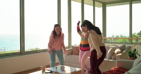 Diverse women dancing and laughing in sunlit coastal living room with ocean view