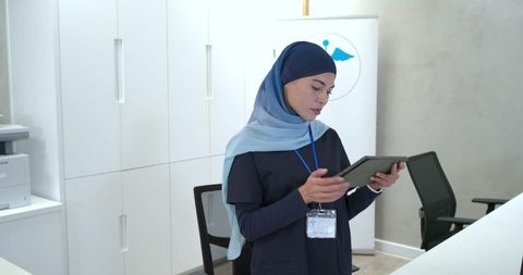 Muslim Female Receptionist Wearing Light Blue Hijab Navy Scrubs Using Tablet in Clinic