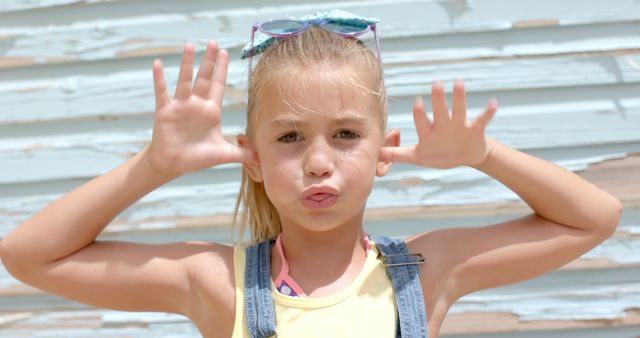 Little girl making silly faces with hands next to cheeks, standing against a light blue wooden wall on a sunny day. She has sunglasses on her head and is wearing casual summer attire. Great for conveying themes of childhood playfulness, outdoor activities, and carefree summer days.