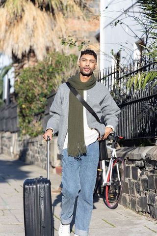 Urban traveler walking with suitcase by stone wall with bicycle in bright sunlight