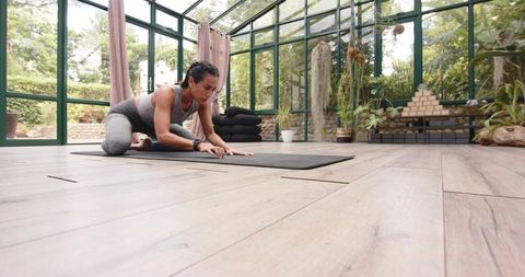 Pacific islander woman stretching in sunroom conservatory on yoga mat with greenery