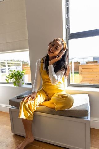 Asian woman enjoying music on window bench in yellow overalls with headphones