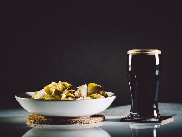 This photo features a glass of dark beer accompanied by a white bowl filled with chips, set on a reflective surface with a dark background. Perfect for utilizations in advertisements related to pubs, bars, snacks, minimalistic dining experiences, or promotions for beer products. Can also be used to illustrate casual dining, nightlife, and social scenarios involving food and drinks.