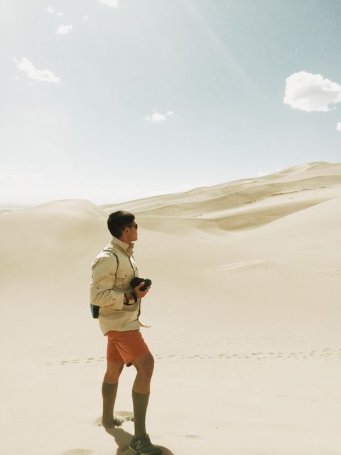 Young man exploring desert dunes during daytime, carrying backpack and holding camera. Useful for themes related to adventure, travel, lifestyle, outdoor activities, and photography.