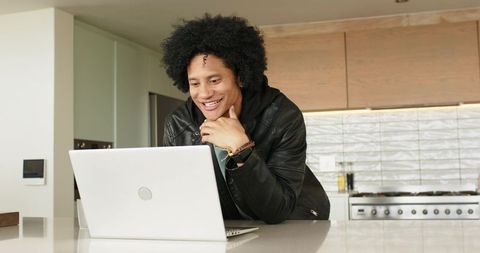 Mid-30s man leaning over white laptop at modern kitchen island smiling while working remotely
