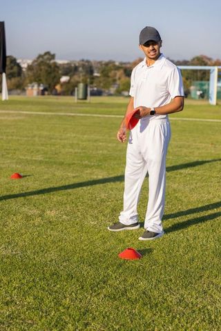 Indian cricketer standing on grass field holding red training cones during outdoor drill