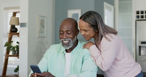 Smiling diverse middle-aged couple sharing smartphone at kitchen table, cozy morning