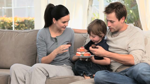 Video depicts a happy family of three enjoying cupcakes in a cozy living room. Mother is giving cakes to her son and husband, showcasing bonding and togetherness. Ideal for use in family, home lifestyle, and parenting-focused content.