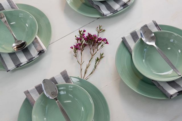 Overhead view of an elegant table setting featuring green plates and bowls, striped napkins, and spoons. A small floral decoration adds a touch of nature to the minimalist and modern arrangement. Ideal for use in articles or advertisements related to dining, home decor, event planning, and hospitality.