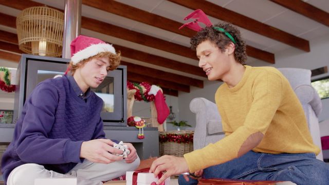 A joyful scene depicting a happy gay couple wrapping Christmas presents by the fireplace in their cozy home. They are wearing festive holiday attire including a Santa hat and reindeer antlers, adding to the seasonal atmosphere. This video can be used to represent love, holiday spirit, and inclusion, ideal for marketing holiday products, lifestyle articles, and inclusivity campaigns.