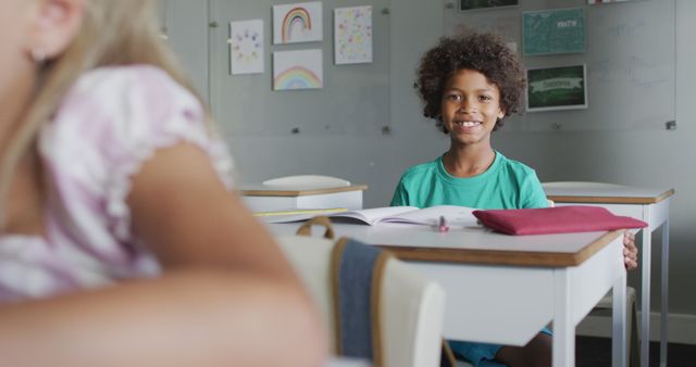 Smiling boy sitting at his desk in a classroom with an open book and colorful artwork displayed on the wall. Perfect for educational themes, back-to-school promotions, children's learning programs, and articles on childhood education and development.