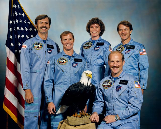 Group of five NASA astronauts in blue uniforms posing with an eagle and an American flag in the background. This photograph, taken in September 1984, features the crew scheduled for the STS-51A mission of the Space Shuttle Discovery. Valuable for historical references, educational materials, or articles on space exploration and NASA's missions.
