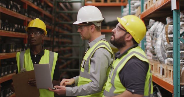 Three warehouse workers in safety gear are standing in an industrial environment. They are discussing logistics next to various storage racks. Two of them are holding a clipboard and laptop, while one worker looks on attentively. This image could be used to illustrate teamwork in warehouse operations, safety protocols, logistics, and inventory management.