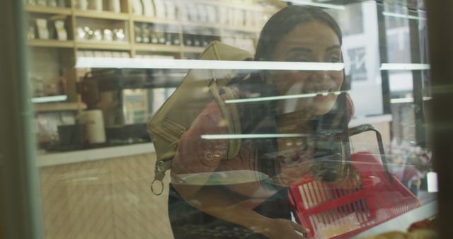 A woman carries a red basket while browsing through items in a supermarket. She smiles as she leans over a display, showing engagement and curiosity. Great for concepts related to grocery shopping, everyday life, and consumer habits. Useful in advertisements for supermarkets, grocery-related articles, and lifestyle blogs.