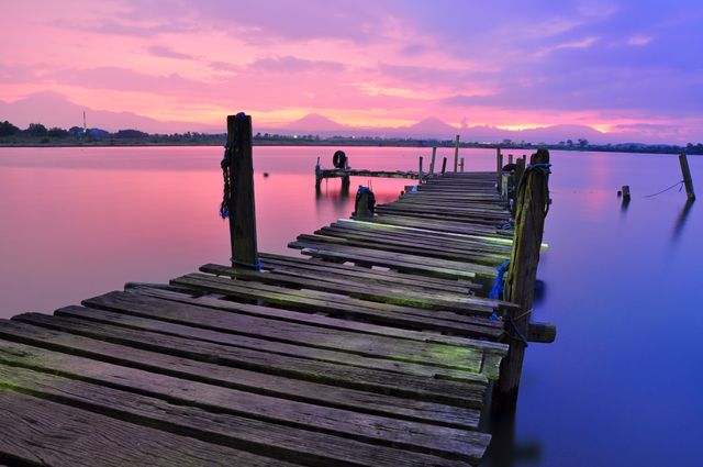 The photo captures a calm and serene old wooden pier with a vibrant sunset sky and reflection in the waters. Useful for concepts related to peace, relaxation, and nature's beauty. Ideal for backgrounds for websites or motivational posters focusing on tranquility and the beauty of nature.