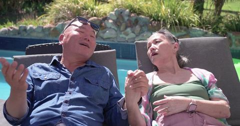 Senior couple holding hands relaxing by poolside loungers in sunny backyard
