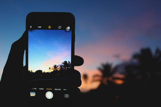 Silhouette holds smartphone taking picture of colorful tropical sunset with palm trees and vibrant sky. Perfect for themes of travel, vacation, photography tips, and social media usage invacation spots.