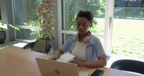 Young african student studying with laptop and notebook by sunlit window