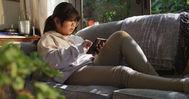 This image features an Asian girl reclining on a couch in a cozy, sunlit living room. She is wearing casual clothing and appears focused on a digital tablet. Ideal for ads or articles about modern living, technology usage, home relaxation, or casual downtime.