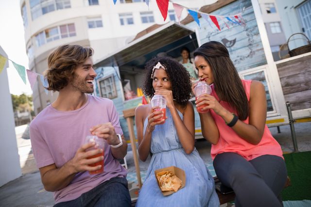 Three friends are enjoying refreshing drinks near a food truck. They are smiling and appear to be having a good time. The setting is urban with colorful decorations, suggesting a casual and fun atmosphere. This image is perfect for promoting social events, food and beverage products, or lifestyle blogs focusing on friendship and outdoor activities.