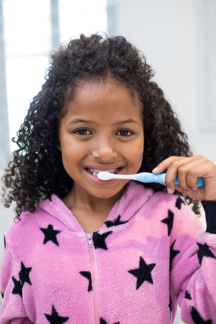 Young girl with curly hair brushing teeth in bathroom, wearing pink pajamas with star pattern. Ideal for promoting children's dental hygiene, morning routines, and healthy habits. Suitable for educational materials, healthcare campaigns, and family lifestyle content.