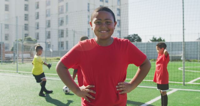 Young boy in red jersey smiling confidently while standing with hands on hips on a soccer field. Other children are playing soccer in the background. Ideal for depicting teamwork, sportsmanship, and outdoor activities for kids. Useful for school projects, sports programs, and community recreation promotions.