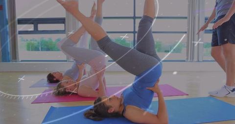 Three women practicing shoulderstand inversions on yoga mats in sunlit studio guided by instructor