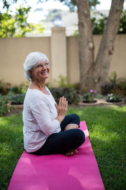 Senior woman sitting on pink yoga mat in park, meditating with hands in prayer position. Ideal for promoting healthy lifestyle, mindfulness, and wellness among elderly. Suitable for articles, blogs, and advertisements focusing on senior fitness, mental health, and outdoor activities.