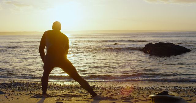 Surfer stretching on beach during sunrise with ocean and horizon in the background. Ideal for fitness, outdoor activity, and morning routine themes.