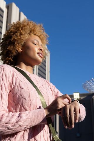 African american woman checking smartwatch in urban sunlight wearing pink knit sweater