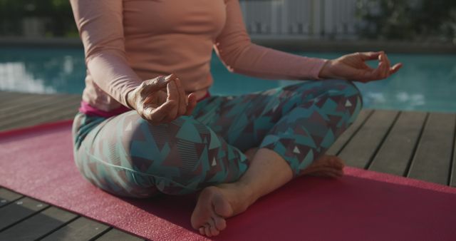 This image shows a woman meditating on a yoga mat by the poolside, dressed in casual wear. Ideal for use in wellness blogs, mental health campaigns, relaxation and fitness promotional materials, and self-care product advertisements. Can also be used to emphasize outdoor or poolside yoga and meditation practices for a tranquil and healthy lifestyle.