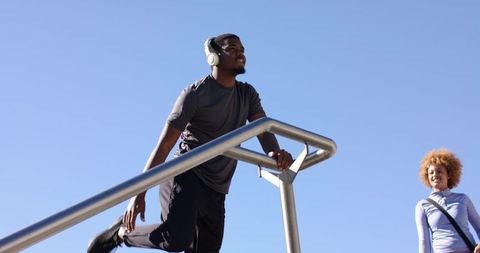 Man stretching on metal rail listening to headphones while woman watching on sunny urban stairs