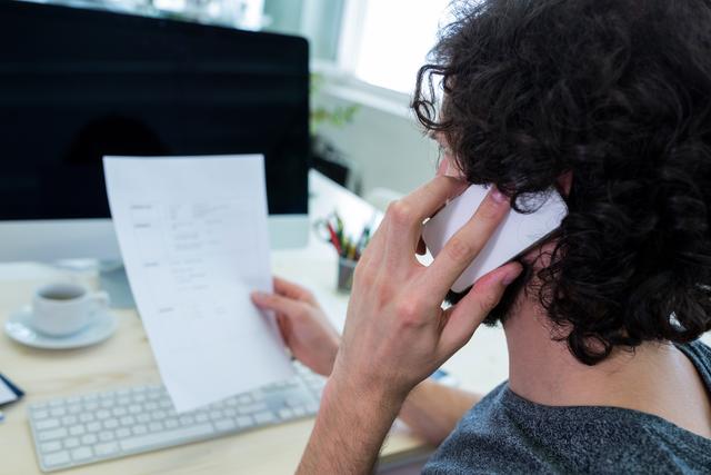 Male graphic designer engaged in a phone conversation while holding a document in a modern office. Ideal for use in articles or advertisements related to creative professions, business communication, remote work, and office environments.
