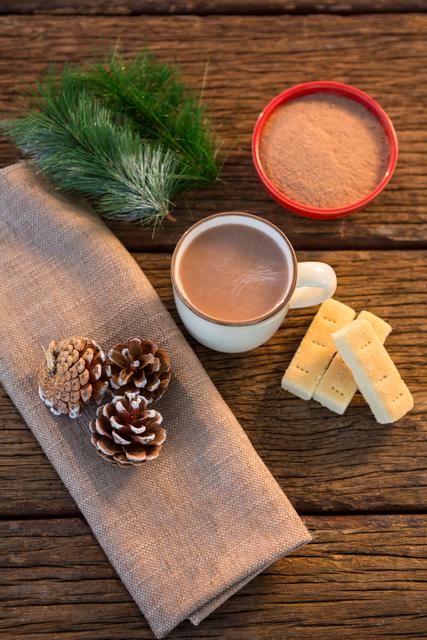 Warm and cozy winter setting featuring a cup of hot coffee, pine cones, fir, sweet treats, and a napkin on a rustic wooden plank. Ideal for use in holiday-themed blogs, cozy winter promotions, festive greeting cards, and beverage advertisements.