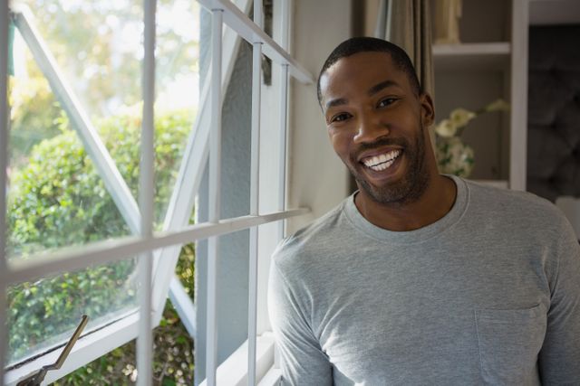 This image depicts a cheerful man standing by a window in a home setting, bathed in natural light. Ideal for use in lifestyle blogs, home decor websites, or advertisements promoting a positive and relaxed domestic atmosphere.