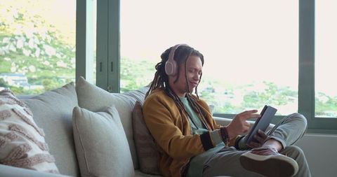 Relaxing man wearing headphones using tablet on modern sofa by large window with scenic view