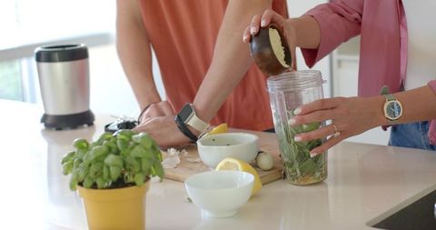 Couple preparing green smoothie with fresh basil lemon and garlic in bright kitchen at counter