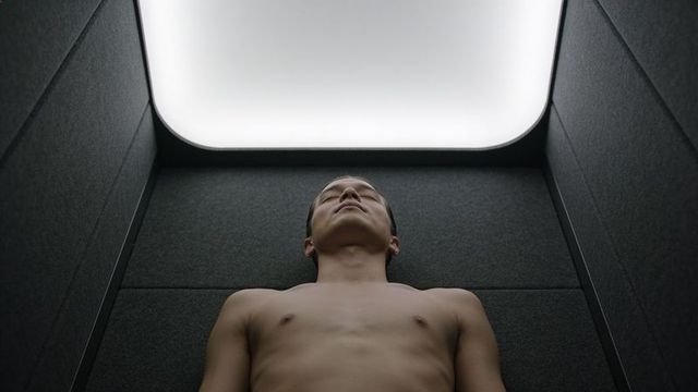 Shirtless asian man reclining inside padded minimalist overhead-lit booth conveying solitude