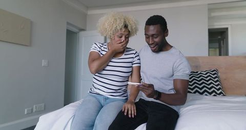 Excited diverse couple discovering positive pregnancy test on bed celebrating pregnancy