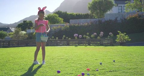 Girl wearing bunny ears holding basket collecting colorful easter eggs on sunny lawn