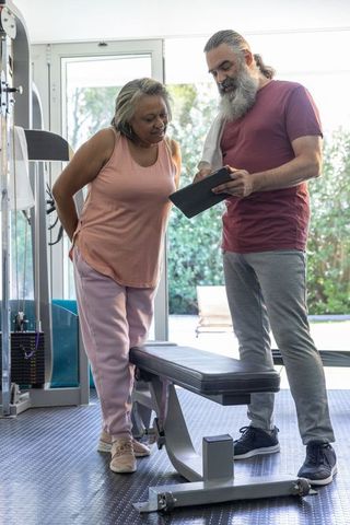 Mature trainer coaching senior woman with tablet beside flat bench in bright home gym